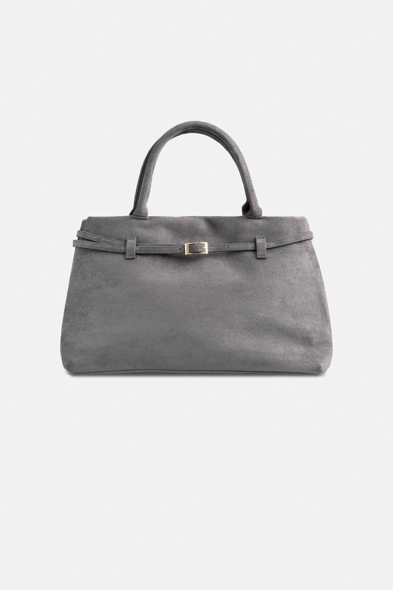 Puede incluir: Un bolso gris con asa superior y detalle de cintur&oacute;n en la parte delantera. El bolso tiene forma rectangular con cierre de solapa y hebilla dorada. El material parece ser ante o un tejido suave similar.