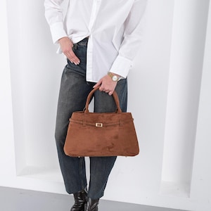 Puede incluir: Un bolso de mano de ante marr&oacute;n con forma rectangular y cierre de hebilla dorada. El bolso tiene dos asas y un detalle de correa en la parte superior. La persona lleva una camisa blanca abotonada y vaqueros oscuros.