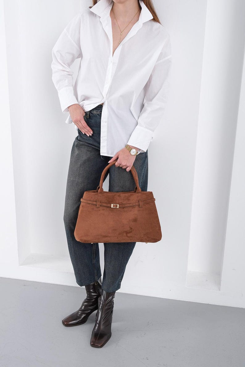 Puede incluir: Un bolso de mano de ante marr&oacute;n con forma rectangular y cierre de hebilla dorada. El bolso tiene dos asas y un detalle de correa en la parte superior. La persona lleva una camisa blanca abotonada y vaqueros oscuros.