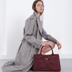 Puede incluir: Un bolso burdeos con forma rectangular y hebilla dorada. El bolso tiene dos asas y un detalle de correa. La persona lleva una gabardina gris.