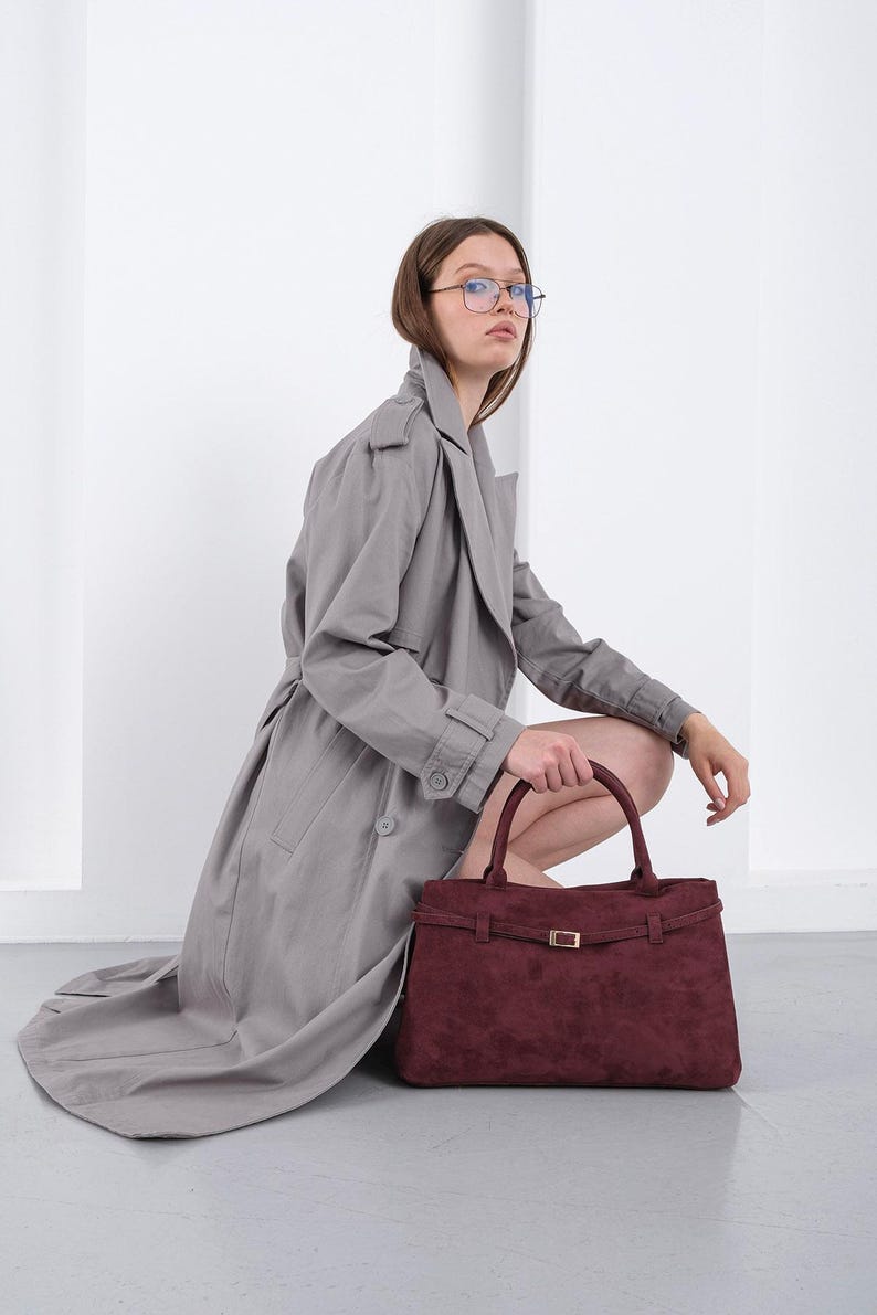 Puede incluir: Un bolso burdeos con forma rectangular y hebilla dorada. El bolso tiene dos asas y un detalle de correa. La persona lleva una gabardina gris.