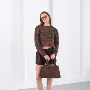 Puede incluir: Una persona viste un su&eacute;ter corto a rayas marrones y verdes, pantalones cortos marrones y botas de gamuza marrones. Sostiene un bolso de mano de gamuza marr&oacute;n con forma rectangular y cierre de hebilla. El fondo es una pared blanca lisa.