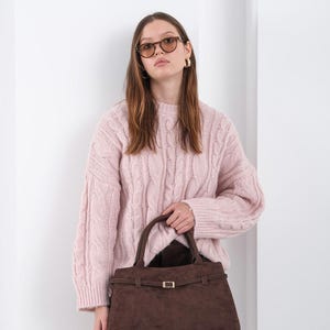 Puede incluir: Una mujer con un su&eacute;ter rosa claro de punto trenzado y gafas de sol marrones sostiene un bolso de mano de gamuza marr&oacute;n. El bolso tiene forma rectangular con un cierre y asa dorados.