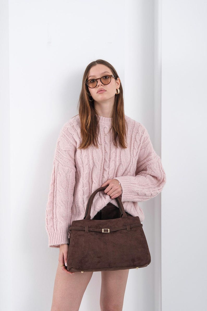 Puede incluir: Una mujer con un su&eacute;ter rosa claro de punto trenzado y gafas de sol marrones sostiene un bolso de mano de gamuza marr&oacute;n. El bolso tiene forma rectangular con un cierre y asa dorados.