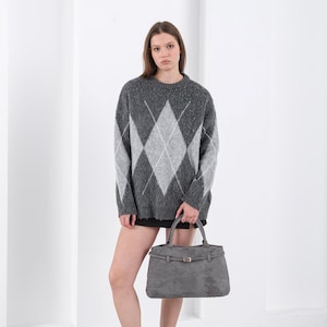 Puede incluir: Un su&eacute;ter gris con estampado de rombos con rombos grises claros y blancos. La persona sostiene un bolso gris de forma rectangular con cierre de hebilla. La persona lleva una falda negra y zapatillas burdeos.