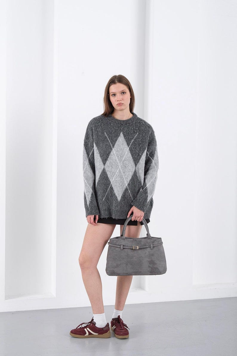 Puede incluir: Un su&eacute;ter gris con estampado de rombos con rombos grises claros y blancos. La persona sostiene un bolso gris de forma rectangular con cierre de hebilla. La persona lleva una falda negra y zapatillas burdeos.