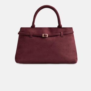 Puede incluir: Bolso de mano burdeos con una silueta estructurada. Cuenta con un asa superior y una fina correa con hebilla dorada. El bolso est&aacute; hecho de un tejido suave y aterciopelado.