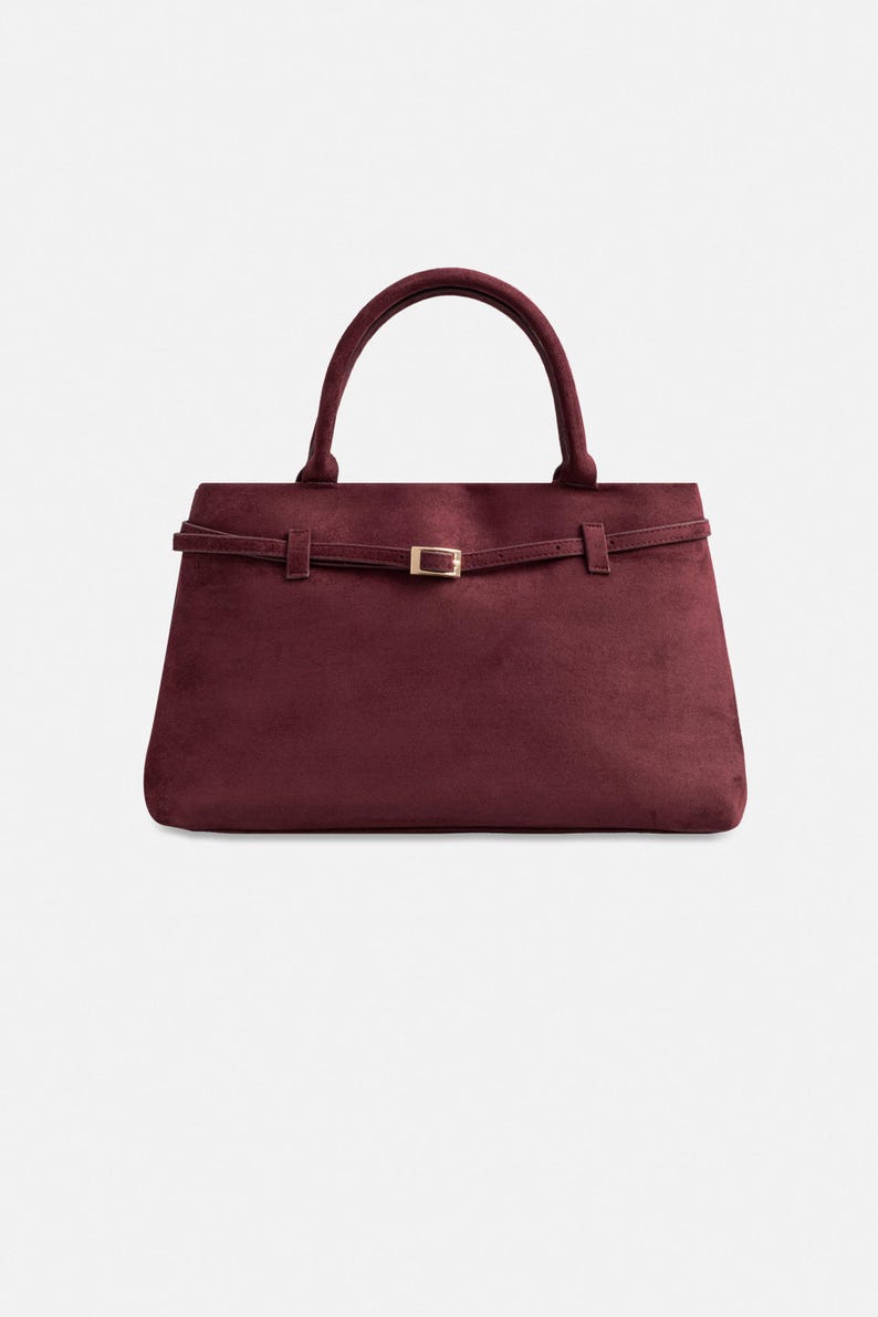 Puede incluir: Bolso de mano burdeos con una silueta estructurada. Cuenta con un asa superior y una fina correa con hebilla dorada. El bolso est&aacute; hecho de un tejido suave y aterciopelado.