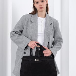 Puede incluir: Un bolso tote de ante negro con forma rectangular y un cierre dorado. El bolso tiene un asa superior y una correa para el hombro. La persona lleva una blazer gris, una camiseta blanca y vaqueros negros.