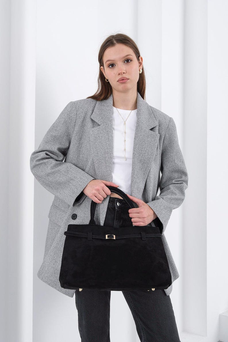 Puede incluir: Un bolso tote de ante negro con forma rectangular y un cierre dorado. El bolso tiene un asa superior y una correa para el hombro. La persona lleva una blazer gris, una camiseta blanca y vaqueros negros.