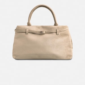 Puede incluir: Bolso de mano de ante beige con asa superior y cierre de solapa. El bolso presenta un detalle de cintur&oacute;n con una peque&ntilde;a hebilla. El dise&ntilde;o general es sencillo y elegante, adecuado para el uso diario.