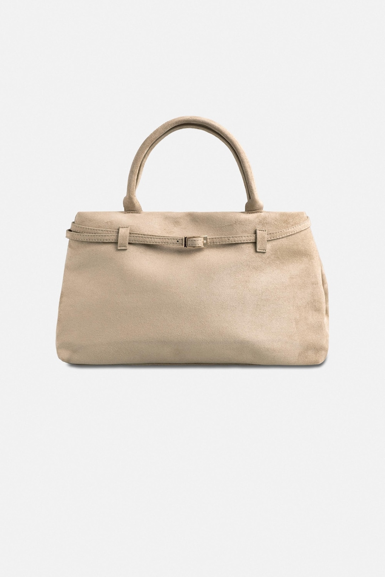 Puede incluir: Bolso de mano de ante beige con asa superior y cierre de solapa. El bolso presenta un detalle de cintur&oacute;n con una peque&ntilde;a hebilla. El dise&ntilde;o general es sencillo y elegante, adecuado para el uso diario.