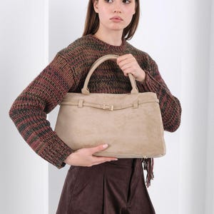 Puede incluir: Bolso de mano de ante beige con forma rectangular y asa superior. El bolso presenta una correa con hebilla dorada. La persona lleva un jersey marr&oacute;n y verde y unos pantalones cortos marrones.