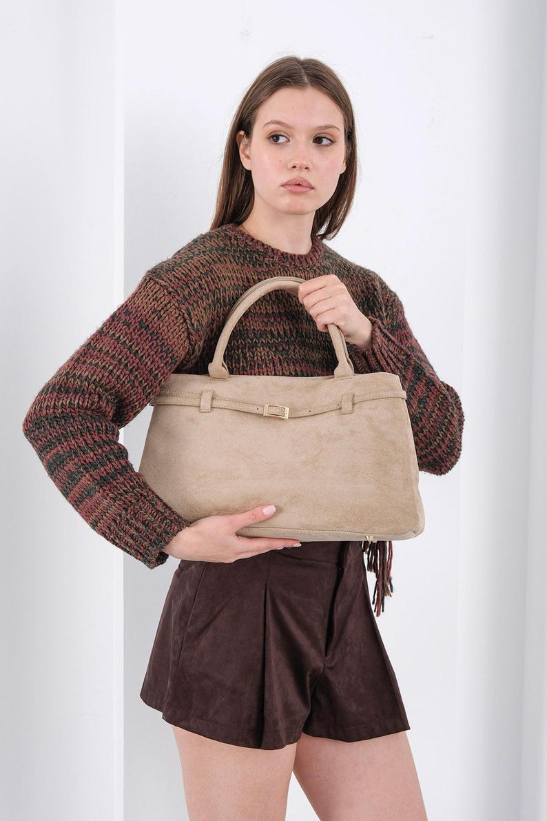 Puede incluir: Bolso de mano de ante beige con forma rectangular y asa superior. El bolso presenta una correa con hebilla dorada. La persona lleva un jersey marr&oacute;n y verde y unos pantalones cortos marrones.