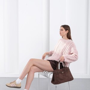 Puede incluir: Un su&eacute;ter rosa claro de punto trenzado y un bolso de mano de ante marr&oacute;n con forma rectangular y cierre de hebilla. La persona lleva pantalones cortos marrones y zapatillas de deporte color canela con rayas rosas.