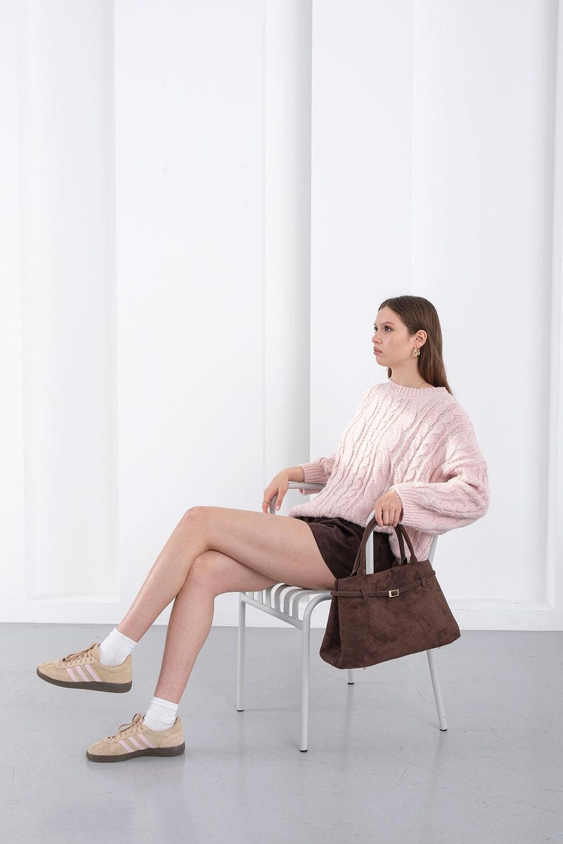 Puede incluir: Un su&eacute;ter rosa claro de punto trenzado y un bolso de mano de ante marr&oacute;n con forma rectangular y cierre de hebilla. La persona lleva pantalones cortos marrones y zapatillas de deporte color canela con rayas rosas.