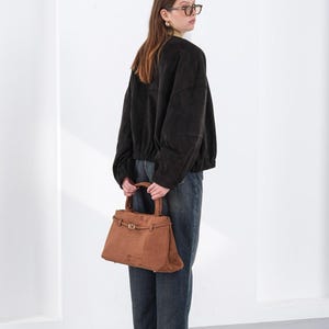 Puede incluir: Un bolso de mano de ante marr&oacute;n con forma rectangular y asa superior. El bolso tiene un cierre de hebilla y un dise&ntilde;o estructurado. La persona de la imagen lleva una chaqueta negra y vaqueros azules.