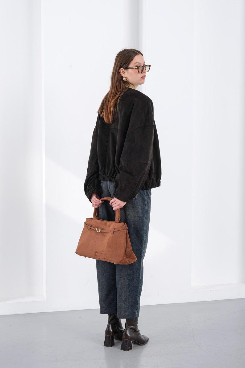 Puede incluir: Un bolso de mano de ante marr&oacute;n con forma rectangular y asa superior. El bolso tiene un cierre de hebilla y un dise&ntilde;o estructurado. La persona de la imagen lleva una chaqueta negra y vaqueros azules.