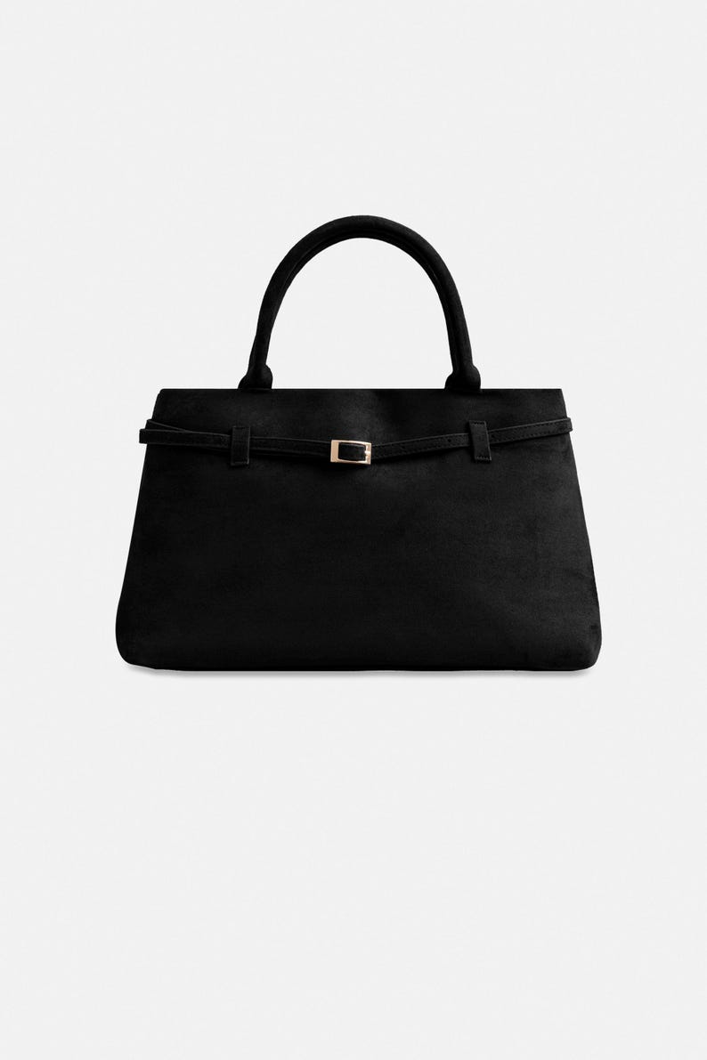 Puede incluir: Bolso de mano negro con forma rectangular y asa curva. El bolso presenta una fina correa con una hebilla dorada. El bolso est&aacute; hecho de un material suave similar al ante.