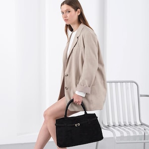 Puede incluir: Una mujer con una blazer beige sostiene un bolso negro con asa superior y hebilla dorada. Viste camisa blanca, pantalones cortos y zapatillas negras con rayas blancas. El bolso es rectangular.