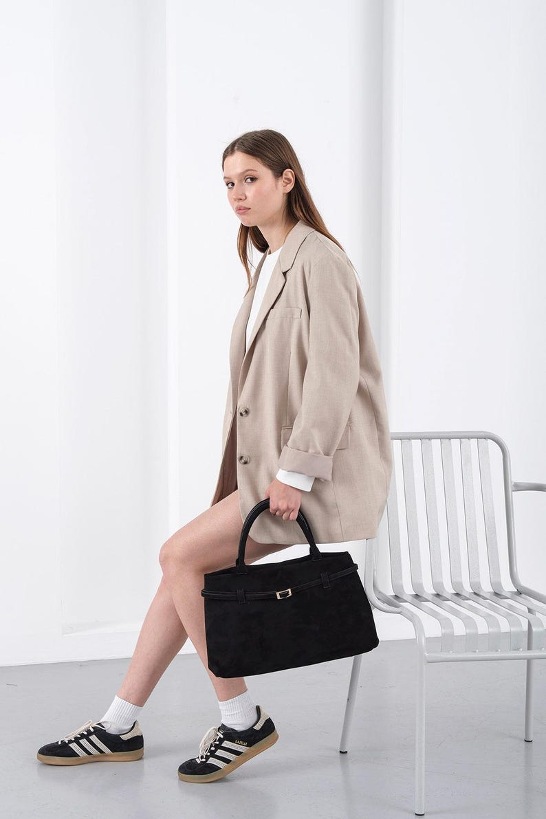 Puede incluir: Una mujer con una blazer beige sostiene un bolso negro con asa superior y hebilla dorada. Viste camisa blanca, pantalones cortos y zapatillas negras con rayas blancas. El bolso es rectangular.