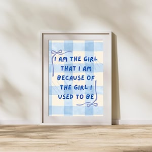 May include: A framed wall art print featuring a light blue and cream gingham check background. The text in dark blue reads, I AM THE GIRL THAT I AM BECAUSE OF THE GIRL I USED TO BE, framed by two light purple hand-drawn ribbon illustrations.