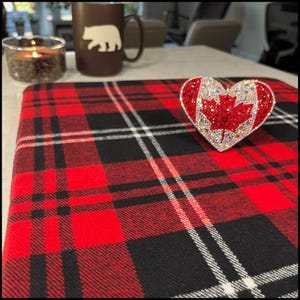May include: A red, black, and white plaid fabric with a heart-shaped decoration. The heart features a Canadian flag design, with a red maple leaf and sparkling embellishments. A brown mug with a bear design is in the background.