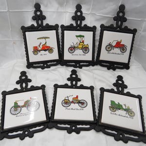 May include: Six vintage-style trivets with black cast iron frames. Each trivet features a white tile with a colorful illustration of an antique car. The cars are in various colors, including red, yellow, and green, with the year of the car printed below.