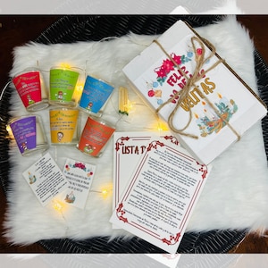 May include: A set of five small glass candle holders with colorful Christmas-themed designs. Each holder features a different image, including a nativity scene, a Christmas tree, and a Santa Claus. The candles are surrounded by a white box with the words "Feliz Navidad" and a brown twine bow. There are also two cards with the words "Lista de deseos" and "Que cada vela que se encienda este año, traiga paz y prosperidad a las familias".