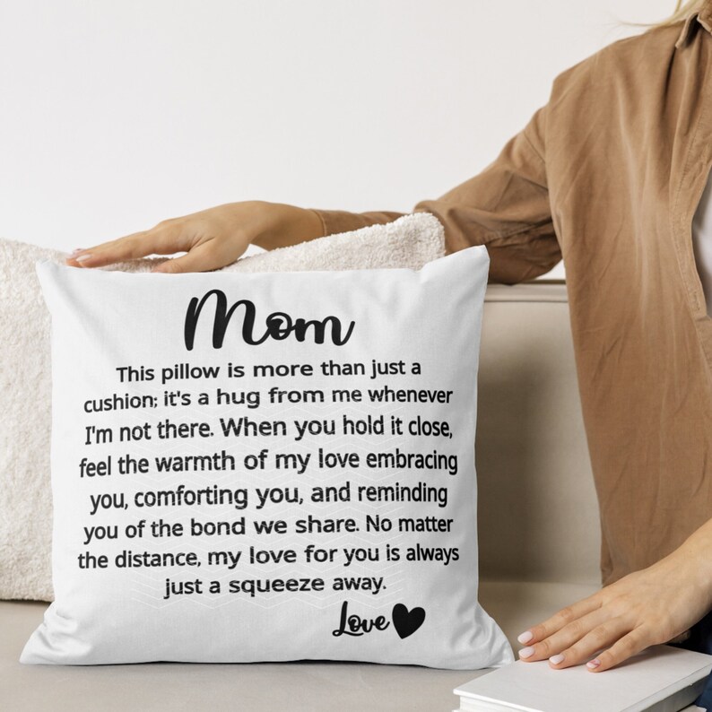 Pode incluir: Travesseiro branco com texto preto que diz "M&atilde;e" e uma mensagem sobre o travesseiro ser um abra&ccedil;o do remetente. O texto tamb&eacute;m inclui a frase "Amor" com um cora&ccedil;&atilde;o preto.
