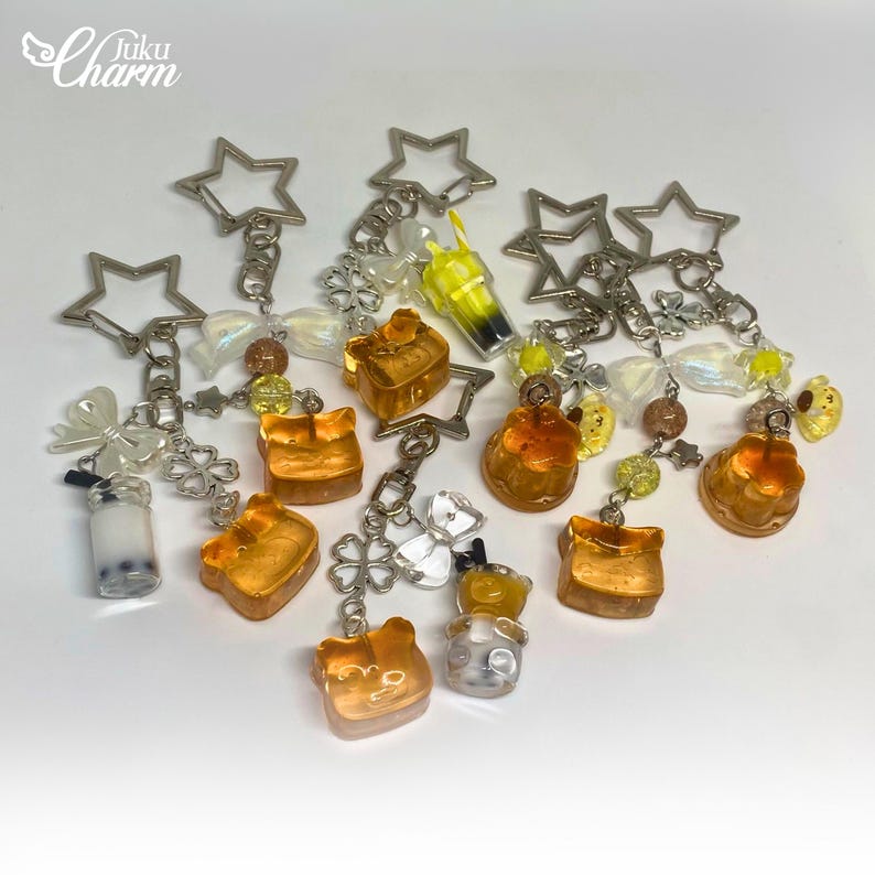 May include: A selection of keyrings featuring food and drink-themed charms. Charms include toast, boba tea, and drinks. Each keyring has a star-shaped clasp and decorative beads. The brand name "Juku Charm" is in the upper left corner.