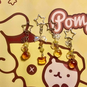 May include: Four decorative keychains with star-shaped tops. Each keychain has a different design, including a bow, a cat, and various beads and charms in shades of yellow, orange, and brown. The keychains are on a yellow background with a cartoon design.