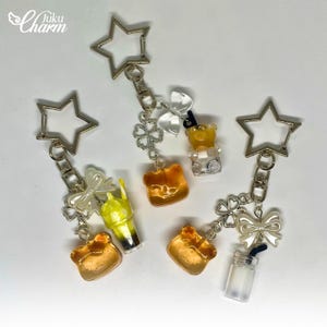 May include: A selection of keyrings with star-shaped clasps. Each keyring features a charm, including a gummy bear, a drink, and a bear-shaped charm. The charms are in shades of yellow, orange, and clear, with silver-tone accents.