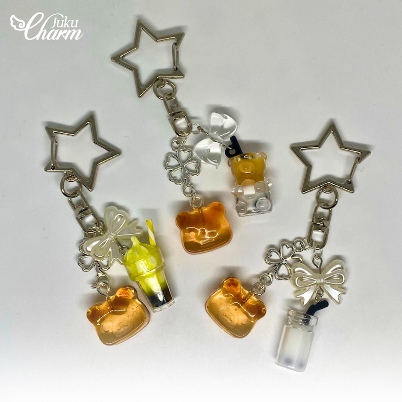 May include: A selection of keyrings with star-shaped clasps. Each keyring features a charm, including a gummy bear, a drink, and a bear-shaped charm. The charms are in shades of yellow, orange, and clear, with silver-tone accents.
