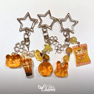 May include: Three keyrings with star-shaped tops and various charms. Charms include a bear, a drink, a four-leaf clover, a butterfly, a pumpkin, a rabbit, and a snack bag. The charms are in shades of orange, yellow, and white.