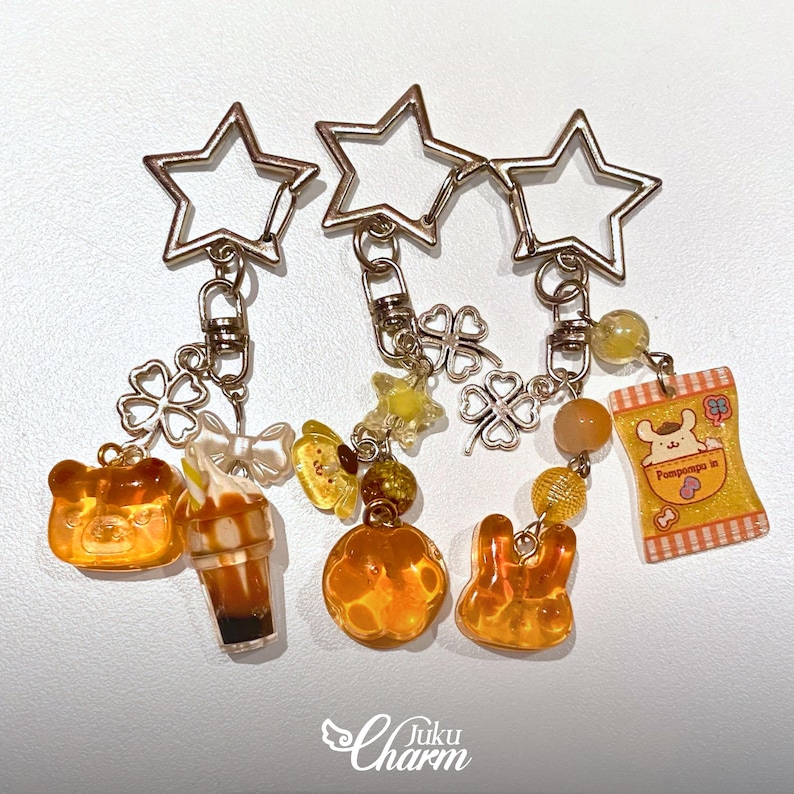 May include: Three keyrings with star-shaped tops and various charms. Charms include a bear, a drink, a four-leaf clover, a butterfly, a pumpkin, a rabbit, and a snack bag. The charms are in shades of orange, yellow, and white.