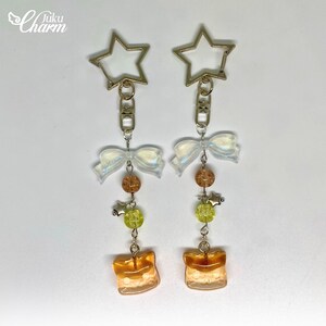 May include: Two keychains with star-shaped clasps. Each keychain features a white iridescent bow, brown and green beads, and a square, amber-coloured charm. The keychains are connected by silver chains.