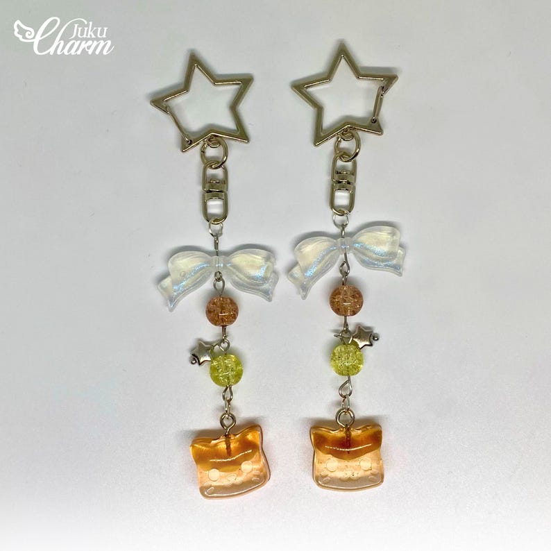 May include: Two keychains with star-shaped clasps. Each keychain features a white iridescent bow, brown and green beads, and a square, amber-coloured charm. The keychains are connected by silver chains.