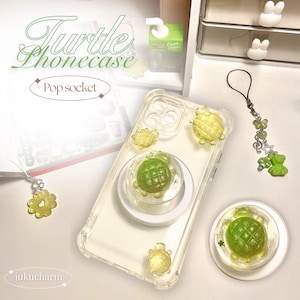 May include: A clear phone case with a pop socket featuring green turtle charms. The phone case has a clear back and sides, and the pop socket is a clear circle with a green turtle charm in the center. The phone case is on a white surface.