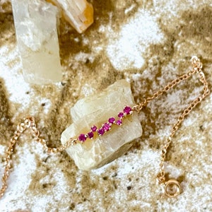 Peut inclure: Un bracelet délicat en chaîne dorée avec une rangée de pierres précieuses roses. Le bracelet est posé sur une surface blanche avec de petits cailloux brun clair.