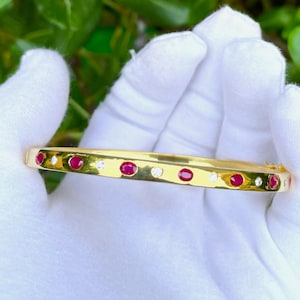 Puede incluir: Una pulsera dorada con piedras preciosas rojas y blancas alternas. La pulsera se sostiene en una mano enguantada sobre un fondo de follaje verde y flores rosas. Las piedras preciosas están espaciadas uniformemente.