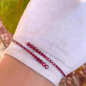 Puede incluir: Una pulsera de estilo bypass con dos filas de piedras preciosas redondas de color rojo vibrante engastadas en una banda de metal plateado pulido. El diseño consiste en una línea continua de piedras que se superponen ligeramente.