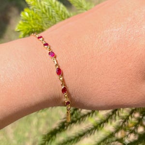 May include: A delicate gold chain bracelet featuring a series of small, oval-shaped, vibrant red gemstones set in individual gold bezels. The bracelet is designed with a thin, polished gold link structure that connects each of the red stones.