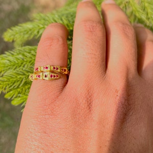 Puede incluir: Un anillo de oro con un diseño geométrico y piedras preciosas rojas. El anillo se lleva en un dedo.