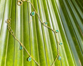 May include: A delicate gold-colored bracelet featuring teardrop-shaped emerald green gemstones. The bracelet is displayed against a backdrop of green palm leaves, highlighting the jewelry's design and color.