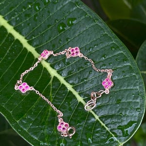 Puede incluir: Un delicado brazalete de cadena dorada con detalles de piedras preciosas rosas. El brazalete presenta una serie de pequeños amuletos de forma cuadrada con piedras rosas engastadas en un marco dorado.