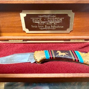 May include: A folding pocket knife with a silver blade and a decorative handle featuring turquoise, red, and black inlays with a gold horse silhouette. The knife rests in a wooden display box with a plaque reading BUCK CUSTOM HAND-MADE BY NAVAJO ARTIST DAVE YELLOWHORSE.