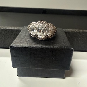 May include: A silver-toned ring featuring three prominent oval-cut clear stones arranged horizontally, surrounded by a halo of smaller pavé-set clear stones. The ring is displayed resting on top of a small, square, black textured jewelry box.