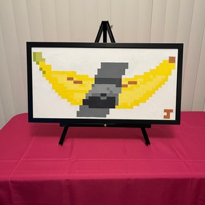 May include: A framed pixel art painting of two yellow bananas with a gray object in the middle, displayed on an easel.