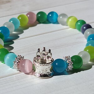 May include: A colorful beaded bracelet with a silver charm shaped like a birthday cake. The beads are green, blue, white, and pink.